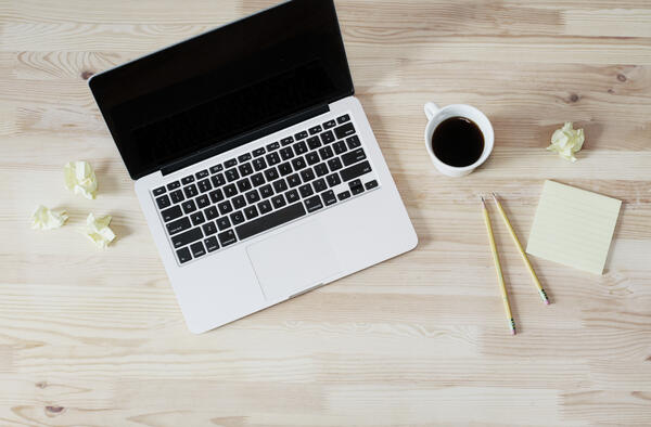 A wooden table with coffee, crumped notes, pencils, and a laptop